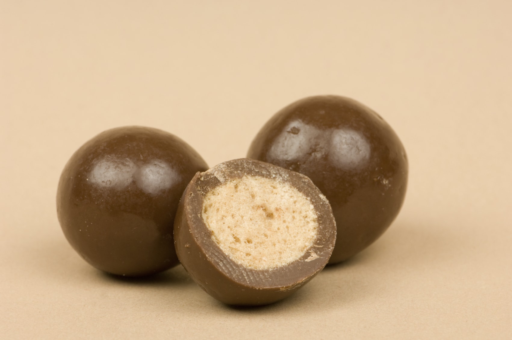 Macro/close-up image of chocolate malted milk balls candy. The front chocolate has been cut in half revealing the hard but airy malt in the middle. On light brown background.